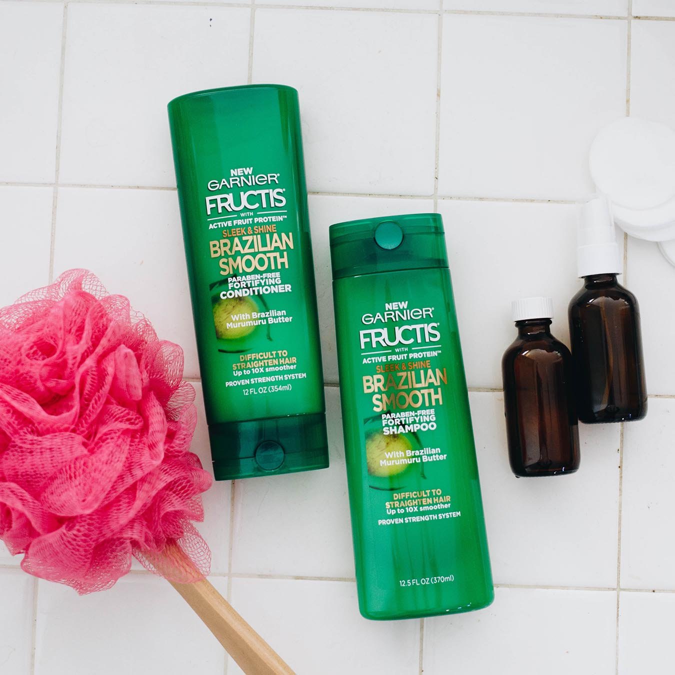Garnier Fructis Brazilian Smooth Shampoo and Brazilian Smooth Conditioner on white tile next to a pink, long-handled bath pouf, to brown dropper bottles, and makeup removal pads.